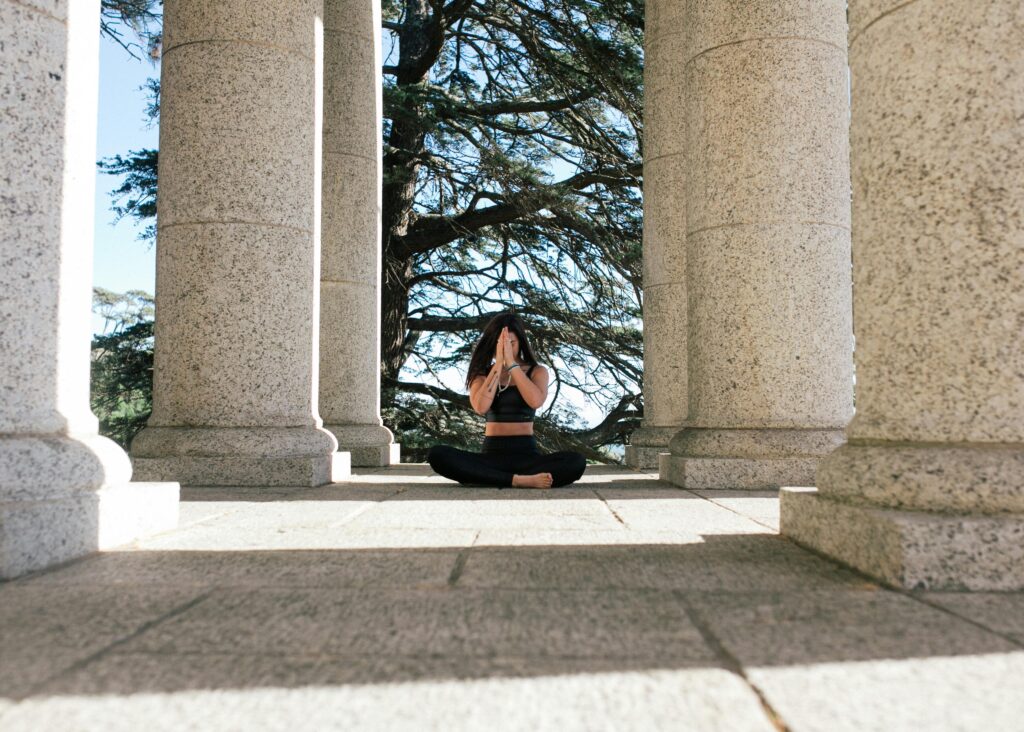 Woman meditating outdoors in a seated yoga pose between stone columns, hands in prayer position, surrounded by trees and natural light.